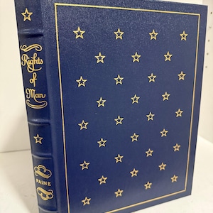 Puede incluir: Un libro de tapa dura azul marino titulado "Rights of Man" de Paine. La portada tiene estrellas doradas y un borde dorado. La columna vertebral muestra el t&iacute;tulo y el nombre del autor en letras doradas.