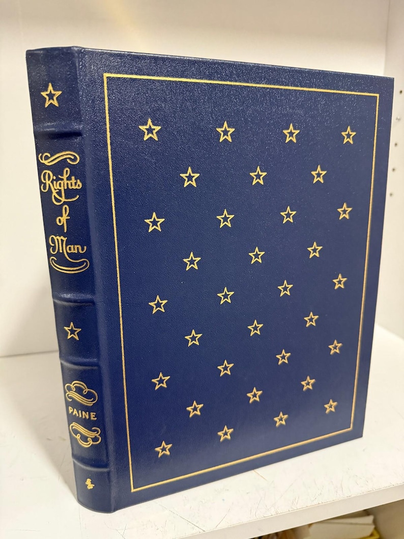 Puede incluir: Un libro de tapa dura azul marino titulado "Rights of Man" de Paine. La portada tiene estrellas doradas y un borde dorado. La columna vertebral muestra el t&iacute;tulo y el nombre del autor en letras doradas.