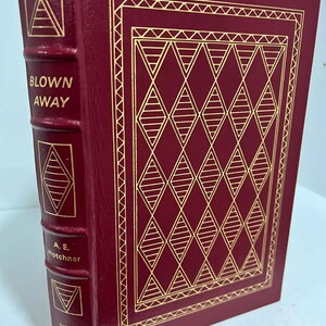 May include: A burgundy leather-bound book with gold lettering and a gold-embossed diamond pattern on the cover. The title is "Blown Away" by A.E. Hotchner. The book is a signed first edition.