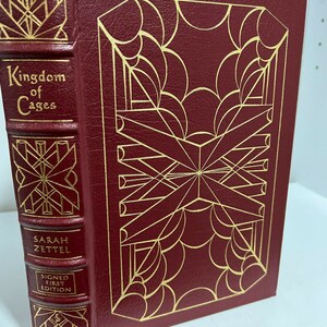 May include: A burgundy leather-bound book with gold lettering and a gold geometric design on the cover. The title of the book is "Kingdom of Cages" by Sarah Zettel. The book is a signed first edition.