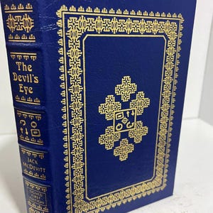 Può includere: Un libro rilegato in pelle blu con lettere dorate e un disegno decorativo dorato sulla copertina. Il titolo del libro è "The Devil's Eye" di Jack McDevitt. Il libro è una prima edizione firmata.