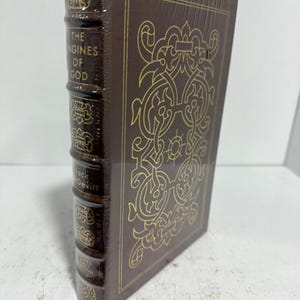 Può includere: Un libro con copertina rigida marrone intitolato "THE ENGINES OF GOD" di Jack McDevitt, avvolto in plastica trasparente. La copertina presenta disegni dorati ornati e un bordo dorato. Il dorso mostra scritte dorate ed elementi decorativi.