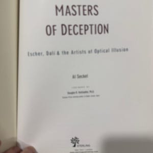 May include: An open book with the title "Masters of Deception" in brown lettering. The subtitle reads "Escher, Dali & the Artists of Optical Illusion." The book's cover is a light beige colour.