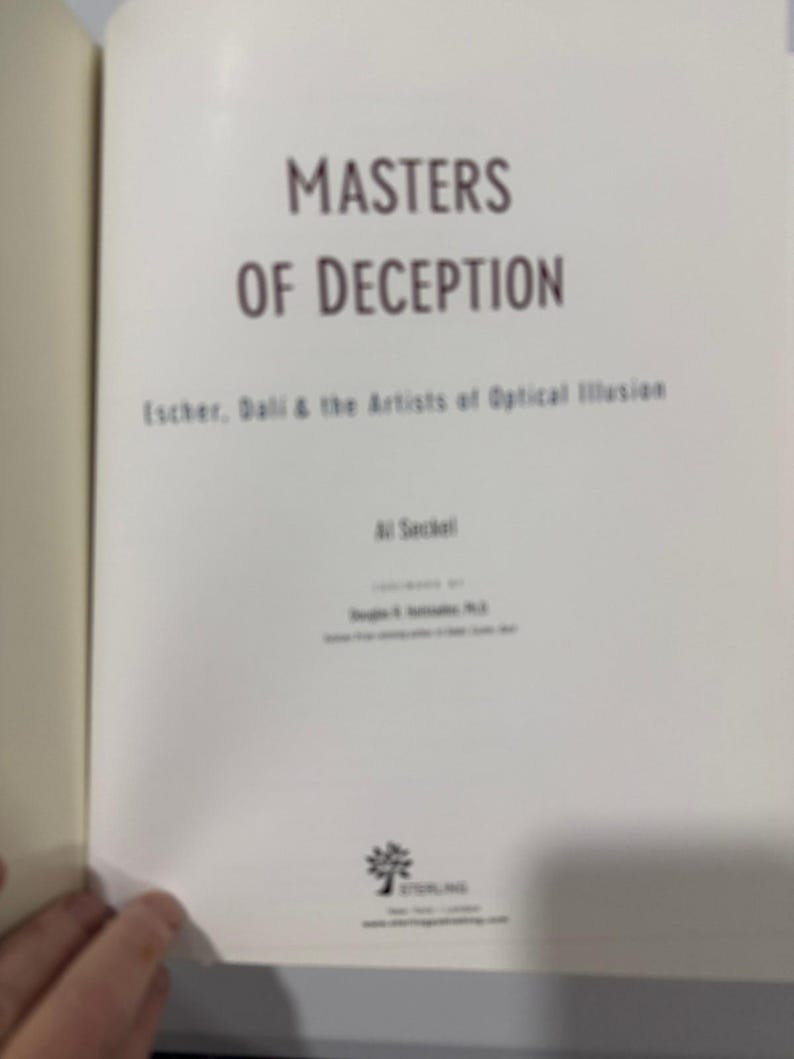 May include: An open book with the title "Masters of Deception" in brown lettering. The subtitle reads "Escher, Dali & the Artists of Optical Illusion." The book's cover is a light beige colour.
