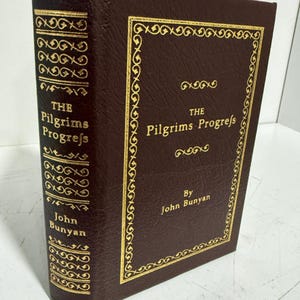 May include: A dark brown hardcover book titled The Pilgrims Progress by John Bunyan. The cover and spine feature gold embossed lettering and decorative border patterns. The book is shown from a front and side angle, displaying the textured finish.