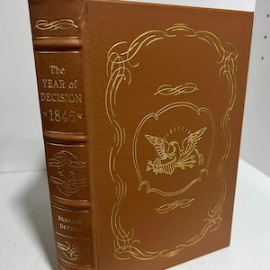 Peut inclure: Un livre relié en cuir marron avec des lettres dorées sur le dos. Le titre est "The Year of Decision 1846" par Bernard Devoto. La couverture présente un motif doré avec un aigle et un bouclier.