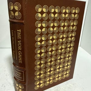 May include: A hardcover book with a brown cover and gold circular designs. The spine reads "THE VOR GAME" and "Lois McMaster Bujold". The book is a signed first edition.