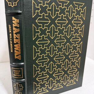 May include: A dark green hardback book titled Mazeway by Jack Williamson. The front cover features a repeating gold geometric pattern. The spine displays the title, author name, and a box with the text Signed First Edition in gold lettering.