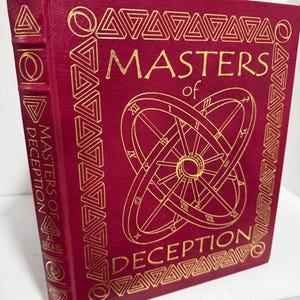 May include: A burgundy hardcover book with gold lettering and geometric patterns. The title "MASTERS of DECEPTION" is prominently displayed. The spine also features the title and author's name. The cover design includes a clock-like illustration.