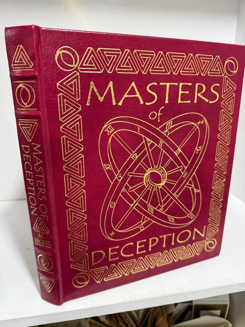 May include: A burgundy hardcover book with gold lettering and geometric patterns. The title "MASTERS of DECEPTION" is prominently displayed. The spine also features the title and author's name. The cover design includes a clock-like illustration.