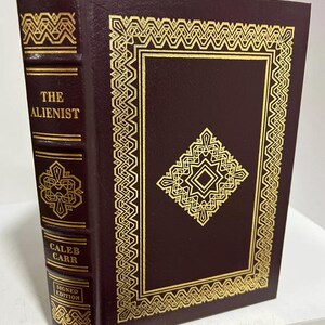 Op de afbeelding: Een bordeauxrood lederen gebonden boek met gouden accenten en een goudkleurig reliëfdesign op de voorzijde. De titel "The Alienist" staat gedrukt op de rug, samen met de naam van de auteur "Caleb Carr" en de woorden "Gesigneerde Editie".