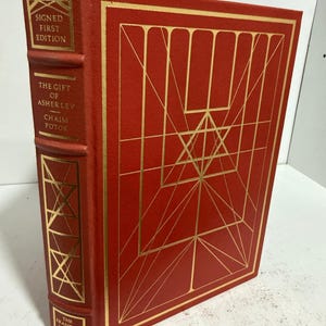 May include: A red hardcover book titled "The Gift of Asher Lev" by Chaim Potok. The book is a signed first edition from The Franklin Library. Gold geometric designs and text are on the cover and spine.