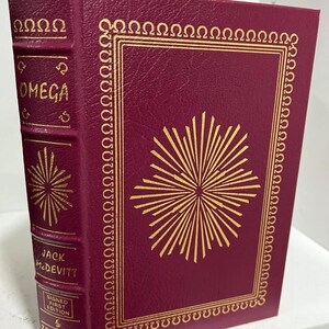 Può includere: Un libro rilegato in pelle bordeaux con dettagli dorati e un design a stella dorata. Il titolo "OMEGA" è stampato sul dorso. Il nome dell'autore "JACK McDEVITT" è stampato sotto il titolo. Il libro è una prima edizione firmata.