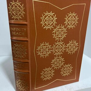 Può includere: Un libro rilegato in pelle marrone con lettere dorate sul dorso. Il titolo è "Infinity Beach" di Jack McDevitt. Il libro è una prima edizione firmata.