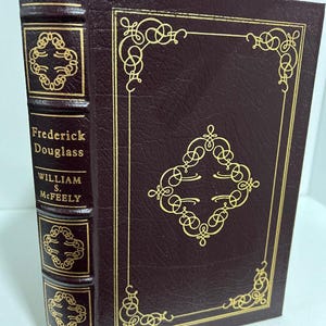 May include: A hardcover book with a burgundy leather cover and gold decorative accents. The book's spine features the title "Frederick Douglass" and the author's name, William S. McFeely. The cover has an ornate gold border and a central design.