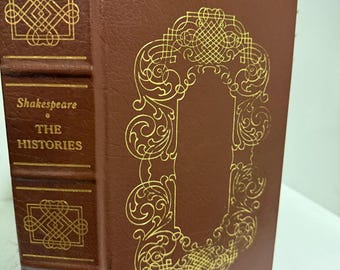 Puede incluir: Un libro de tapa dura marrón titulado Shakespeare THE HISTORIES con patrones de filigrana dorados en relieve en la portada y el lomo. El lomo presenta bandas horizontales en relieve y diseños geométricos repetitivos en tinta dorada.