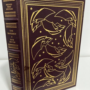 May include: A burgundy leather-bound book with gold lettering and a gold design on the cover. The title is "The Paperboy" by Pete Dexter. The book is a signed first edition and part of The Franklin Library.