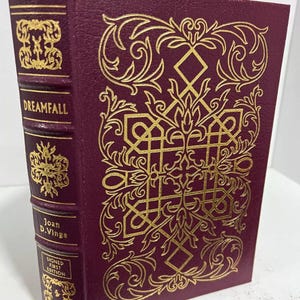 May include: A burgundy leather-bound book with gold lettering and intricate gold designs on the cover. The title "Dreamfall" and the author "Joan D. Vinge" are visible. The book is a signed first edition.
