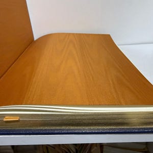 Puede incluir: Libro abierto con una cubierta de imitaci&oacute;n madera y p&aacute;ginas con bordes dorados. El libro tiene una lomo azul y un marcador dorado. Las p&aacute;ginas son blancas y la cubierta es de color marr&oacute;n c&aacute;lido.