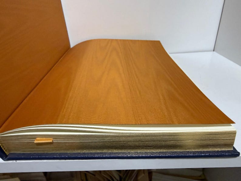 Puede incluir: Libro abierto con una cubierta de imitaci&oacute;n madera y p&aacute;ginas con bordes dorados. El libro tiene una lomo azul y un marcador dorado. Las p&aacute;ginas son blancas y la cubierta es de color marr&oacute;n c&aacute;lido.