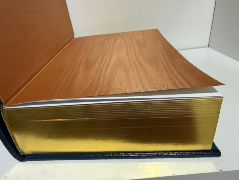 Puede incluir: Un libro abierto con bordes dorados y una cubierta azul oscuro. Las p&aacute;ginas son blancas y la cubierta interior es de color marr&oacute;n c&aacute;lido con un patr&oacute;n de vetas de madera. El libro est&aacute; sobre una superficie blanca.