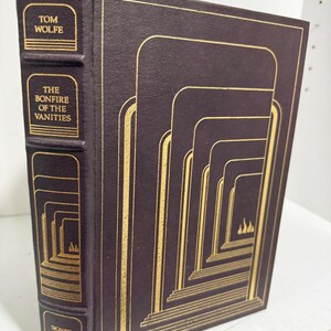 May include: A dark purple hardcover book titled The Bonfire of the Vanities by Tom Wolfe. The cover and spine feature gold foil stamping with a repeating archway design and small flame icons. The spine text reads Tom Wolfe, The Bonfire of the Vanities, and Signed First Edition.