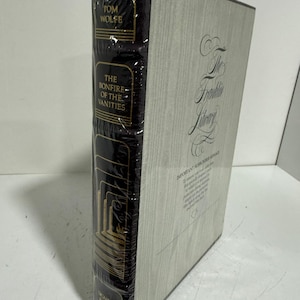 May include: A hardcover book titled The Bonfire of the Vanities by Tom Wolfe, published by The Franklin Library. The book has a black spine with gold lettering and a light gray cover with wood grain texture, all encased in clear plastic shrink wrap.