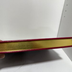 May include: A closed book with a gold-gilded edge and a red cover. The book's spine is visible, showing the gold-coloured pages and the red cover's edge. The book is held by a hand.