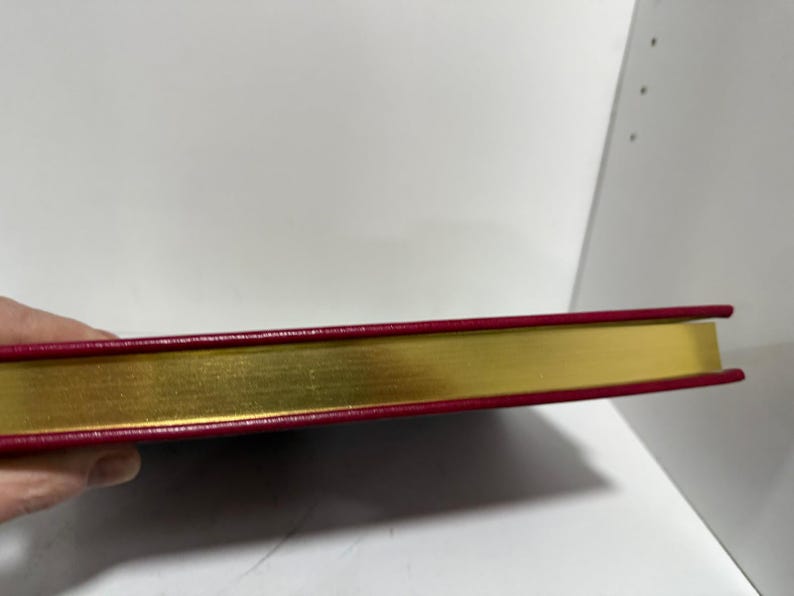 May include: A closed book with a gold-gilded edge and a red cover. The book's spine is visible, showing the gold-coloured pages and the red cover's edge. The book is held by a hand.