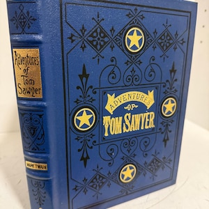 Puede incluir: Un libro de tapa dura azul titulado Adventures of Tom Sawyer de Mark Twain. La portada presenta patrones de filigrana negros, cuatro estrellas amarillas en círculos y una banda amarilla con el título. El lomo incluye el título y el autor.