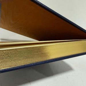 Puede incluir: Un libro parcialmente abierto con bordes dorados y una cubierta azul marino. El interior de la cubierta es de color marr&oacute;n dorado c&aacute;lido. Se ve un peque&ntilde;o marcador amarillo.