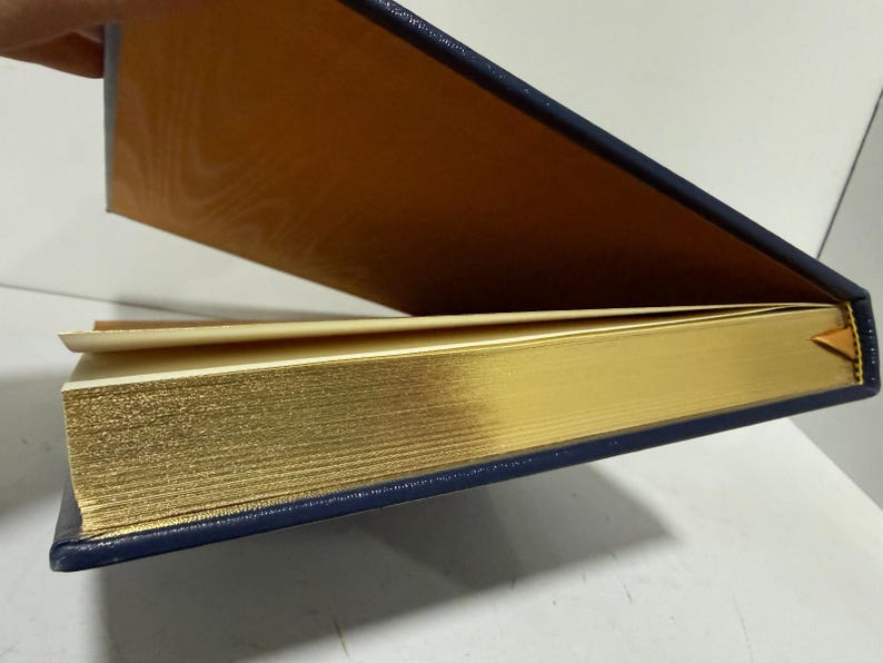Puede incluir: Un libro parcialmente abierto con bordes dorados y una cubierta azul marino. El interior de la cubierta es de color marr&oacute;n dorado c&aacute;lido. Se ve un peque&ntilde;o marcador amarillo.