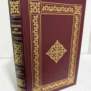 Pode incluir: Um livro de capa dura bordô intitulado "Deadlines and Datelines" de Dan Rather. O livro tem detalhes dourados e texto. A lombada diz "Signed First Edition".