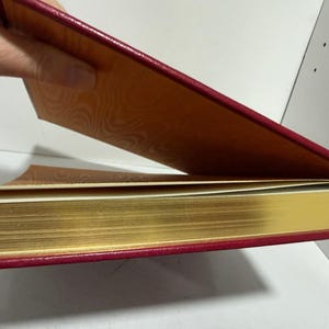 May include: A hardback book with a red cover and gold-gilded pages. The inside cover is patterned in orange. The book is partially open, showing the gold-edged pages and the patterned interior.