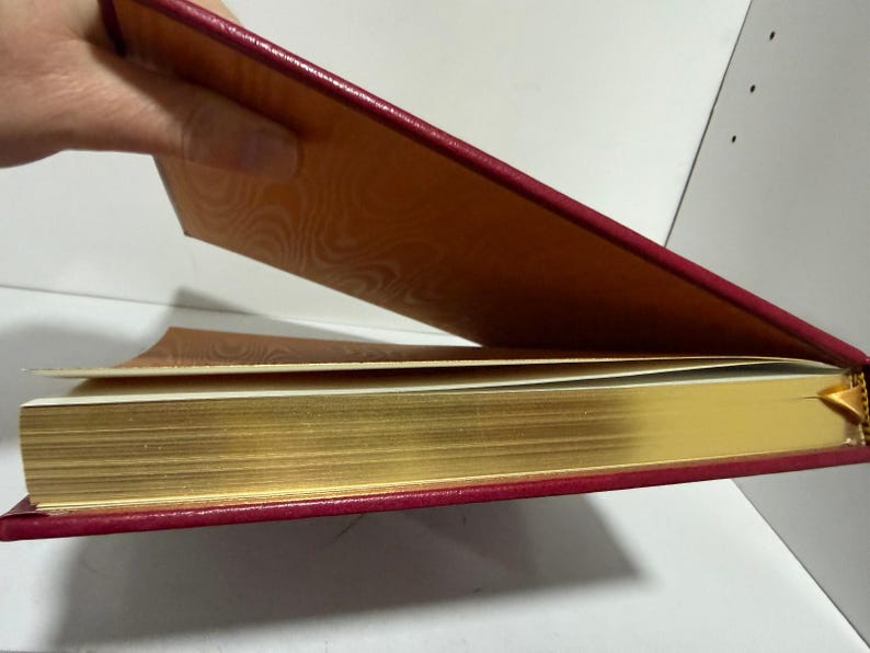 May include: A hardback book with a red cover and gold-gilded pages. The inside cover is patterned in orange. The book is partially open, showing the gold-edged pages and the patterned interior.