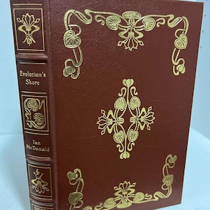 Może przedstawiać: Twarda oprawa książki "Evolution's Shore" autorstwa Iana McDonalda. Książka ma bordową okładkę ze złotymi, kwiatowymi zdobieniami. Grzbiet zawiera tytuł, autora i napis "Signed First Edition".
