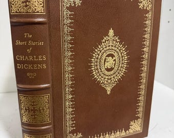 Puede incluir: Un libro encuadernado en cuero marrón titulado The Short Stories of Charles Dickens. La cubierta y el lomo presentan intrincados estampados en pan de oro, incluyendo bordes decorativos y un diseño de medallón central en la portada.