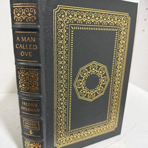 May include: A hardcover book titled A Man Called Ove by Fredrik Backman, featuring a dark charcoal cover with intricate gold foil filigree patterns and an octagonal design on the front, with the title and author name printed on the spine.