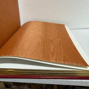 May include: An open book with a red cover and gold-gilded pages. The top page is a light brown wood veneer, with the grain visible. The book is on a white shelf.