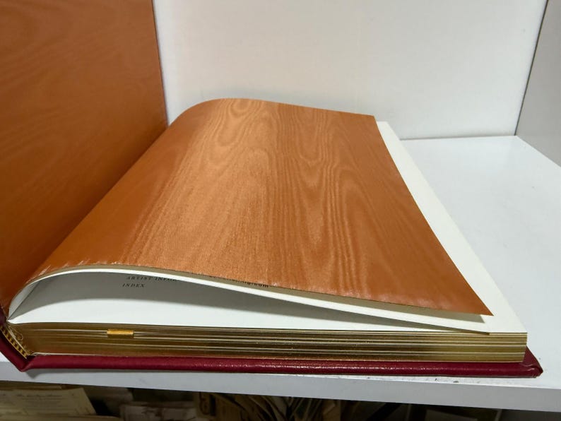 May include: An open book with a red cover and gold-gilded pages. The top page is a light brown wood veneer, with the grain visible. The book is on a white shelf.