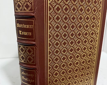 Könnte beinhalten: Ein Hardcover-Buch mit dem Titel "Barchester Towers" von Anthony Trollope. Das Buch hat einen dunkelroten Einband mit goldenen geometrischen Mustern und goldener Schrift. Der Buchrücken weist ebenfalls goldene geometrische Designs und den Titel auf.