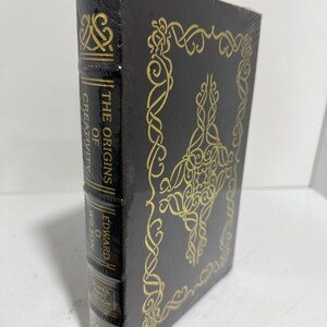 May include: A black hardcover book titled The Origins of Creativity by Edward O. Wilson, wrapped in clear plastic. The cover and spine feature intricate gold foil Celtic-style knotwork patterns. The spine text reads The Origins of Creativity, Edward O. Wilson, and Signed First Edition.