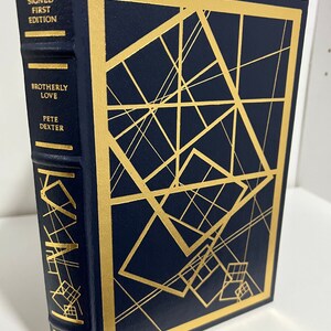 May include: A navy blue hardcover book with gold lettering and a gold geometric design on the cover. The title of the book is "Brotherly Love" by Pete Dexter. The book is a signed first edition and is part of The Franklin Library.