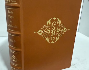 Puede incluir: Un libro de tapa dura marrón titulado "Early Days in California" de Field. La portada presenta detalles decorativos y texto dorados. El lomo muestra el título y el nombre del autor en dorado. El libro se exhibe sobre una superficie blanca.