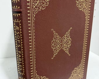 Puede incluir: Un libro de tapa dura color granate titulado Aesop's Fables con detalles de filigrana en lámina dorada en la portada y el lomo. La portada presenta un motivo decorativo central y un borde de símbolos en forma de cruz, con el título en el lomo.