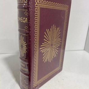 Può includere: Un libro rilegato in bordeaux con lettere dorate e un disegno dorato sulla copertina. Il titolo del libro è "Omega" e l'autore è "Jack Devitte". Il libro è una prima edizione firmata.