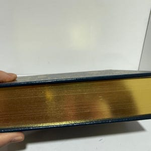 Puede incluir: Un libro de tapa dura con bordes dorados y una cubierta azul oscuro. Las p&aacute;ginas del libro son de color dorado brillante y la cubierta es azul oscuro con un patr&oacute;n sutil. El libro est&aacute; sostenido por una mano.