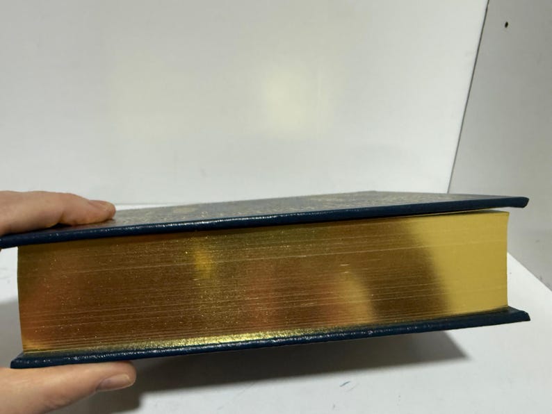 Puede incluir: Un libro de tapa dura con bordes dorados y una cubierta azul oscuro. Las p&aacute;ginas del libro son de color dorado brillante y la cubierta es azul oscuro con un patr&oacute;n sutil. El libro est&aacute; sostenido por una mano.