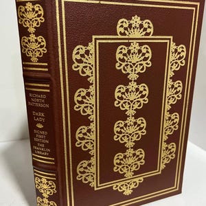 May include: A burgundy leather-bound book with gold accents and a gold-embossed design. The title "Dark Lady" is printed on the spine, along with the author's name "Richard North Patterson". The book is a signed first edition from The Franklin Library.