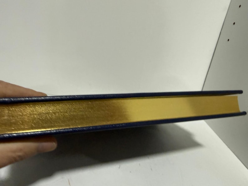 Puede incluir: Un libro con una cubierta azul marino y bordes dorados. Las p&aacute;ginas del libro son doradas y la cubierta es de color azul oscuro. El libro est&aacute; sostenido por una mano.